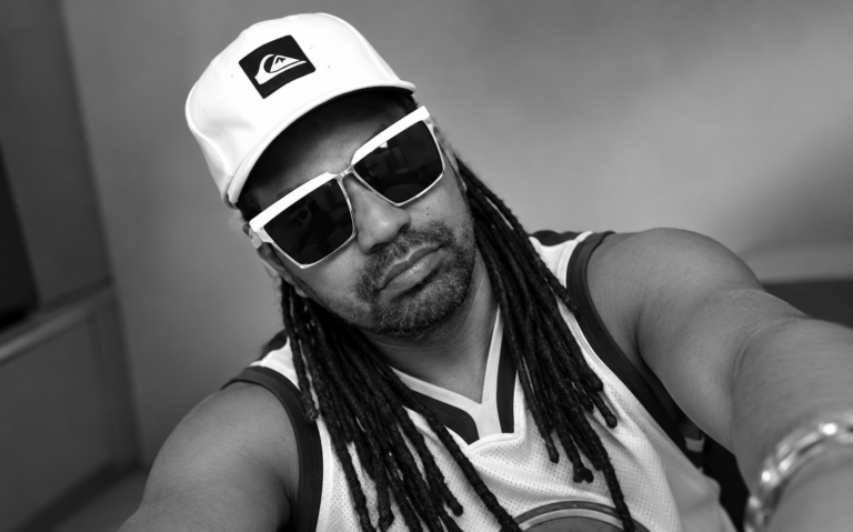 Músico e produtor cultural Edu Bizu usando boné branco, óculos escuros e cabelos em dreadlocks, em fotografia preto e branco.