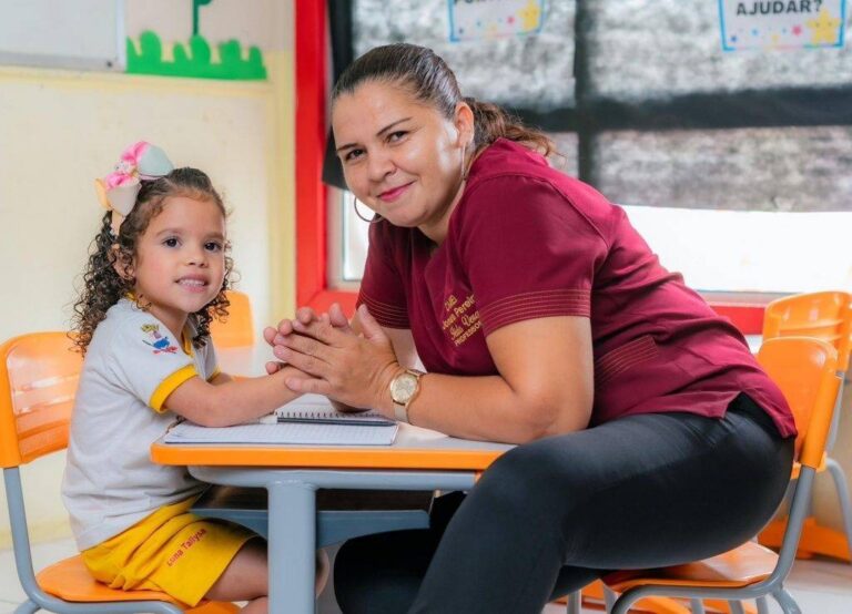 Profissional da educação com blusa vinho acompanha criança sentada à mesa em sala de aula, com cadeiras escolares laranjas.