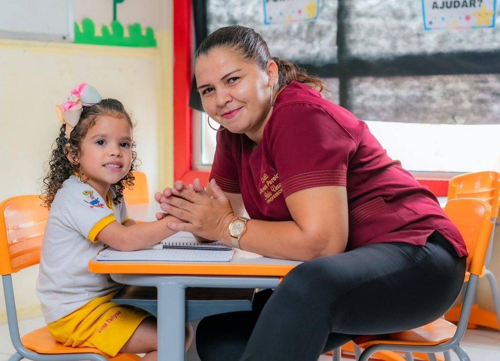 Profissional da educação com blusa vinho acompanha criança sentada à mesa em sala de aula, com cadeiras escolares laranjas.