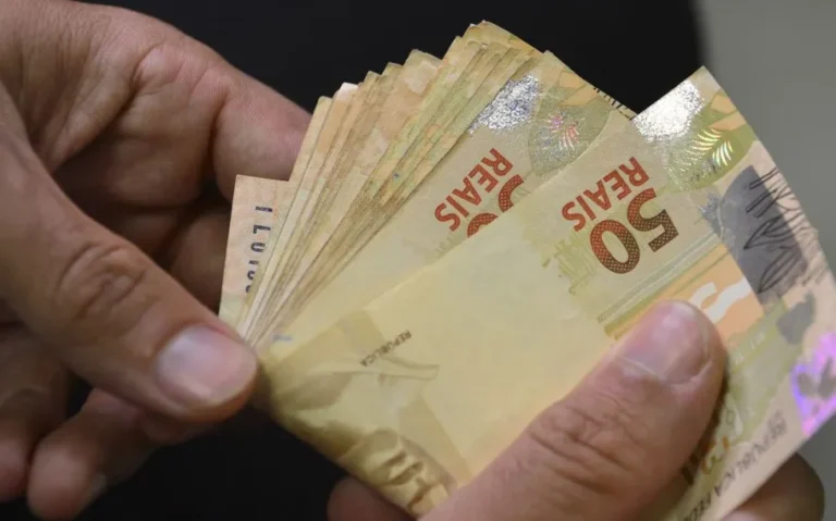Duas mãos seguram várias cédulas de 50 reais abertas em leque, em primeiro plano, sobre fundo escuro.