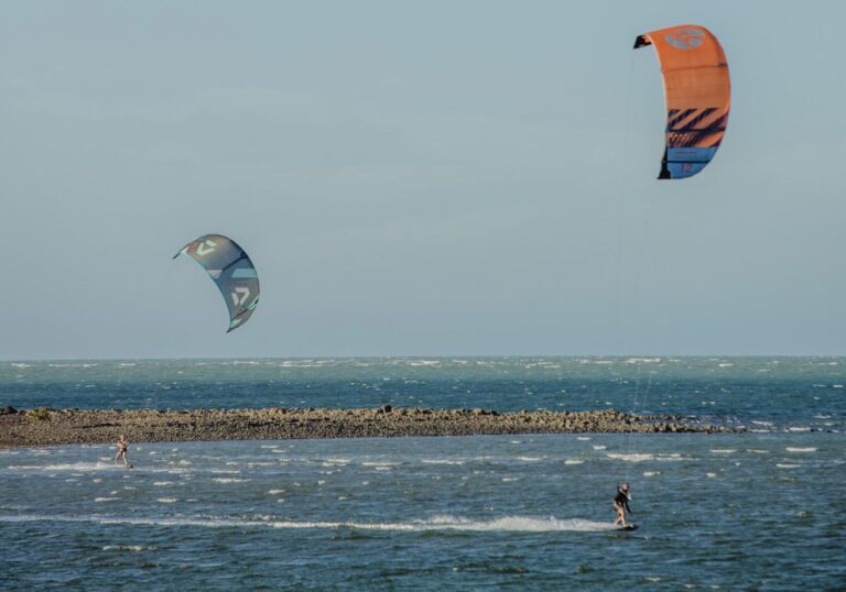Dois praticantes de kitesurfe deslizam sobre o mar, com pipas coloridas no céu e um quebra-mar ao fundo.