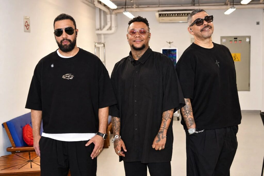 Três homens vestidos de preto, usando óculos, posam juntos para uma fotografia em ambiente interno.