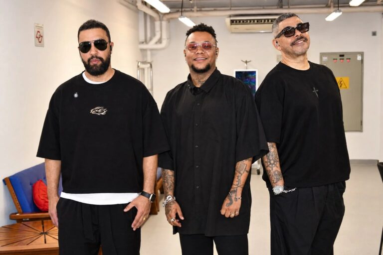 Três homens vestidos de preto, usando óculos, posam juntos para uma fotografia em ambiente interno.