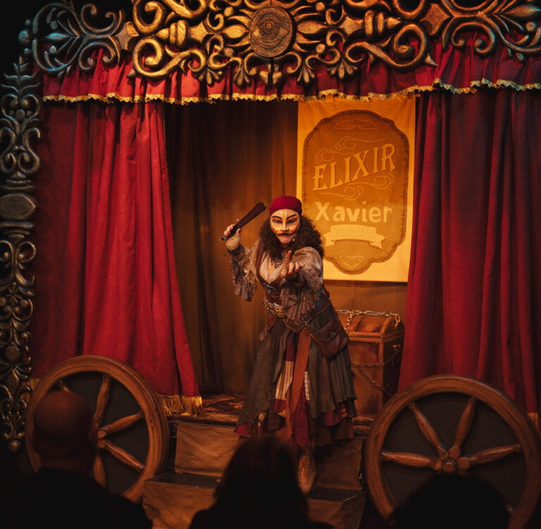 Artista com figurino e maquiagem cênica em palco de cortinas vermelhas, diante de cenário com o nome Elixir Xavier.
