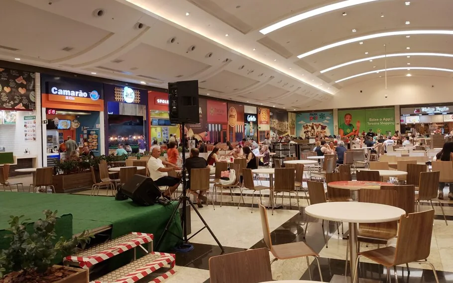 Praça de alimentação de shopping com mesas, cadeiras, clientes e estabelecimentos de comida ao fundo.
