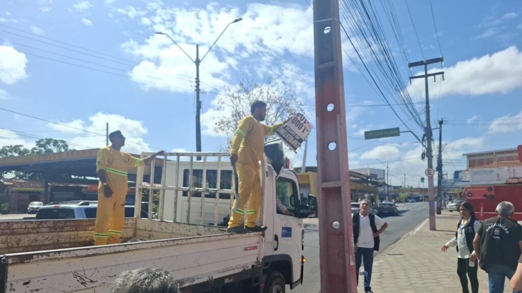 Trabalhador de uniforme amarelo segura uma placa publicitária sobre um caminhão, ao lado de um poste, em rua com calçada, veículos e fiação elétrica.
