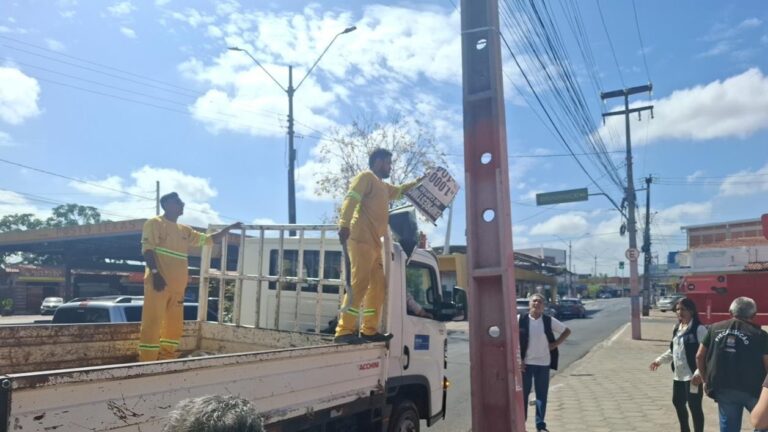 Trabalhador de uniforme amarelo segura uma placa publicitária sobre um caminhão, ao lado de um poste, em rua com calçada, veículos e fiação elétrica.