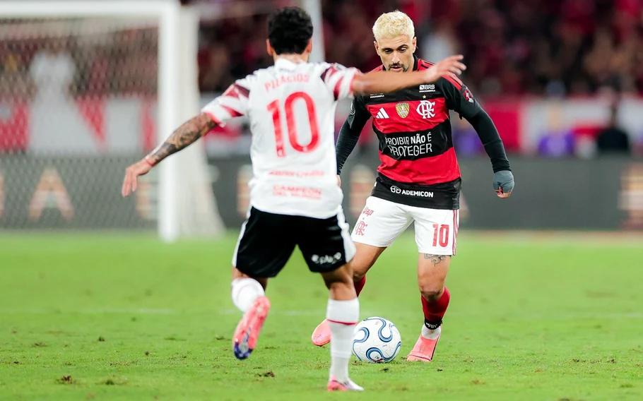 Jogador do Flamengo com uniforme rubro-negro conduz a bola diante de adversário do Estudiantes, de camisa branca número 10.