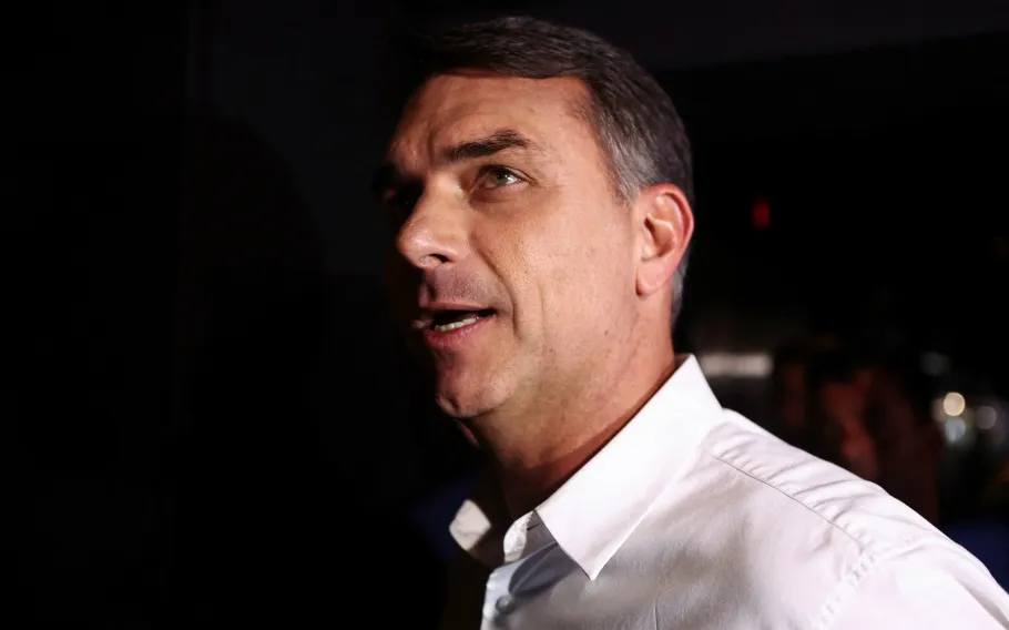 Flávio Bolsonaro de perfil, usando camisa branca, fala em ambiente escuro