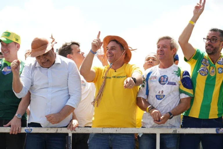 Flávio Bolsonaro, de camisa amarela e chapéu de couro, sorri e gesticula ao lado de outros políticos e apoiadores sobre um trio elétrico.