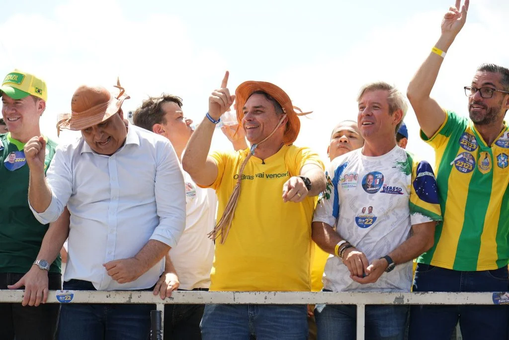 Flávio Bolsonaro, de camisa amarela e chapéu de couro, sorri e gesticula ao lado de outros políticos e apoiadores sobre um trio elétrico.