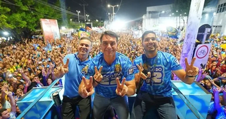 Flávio Bolsonaro e dois aliados, vestidos de azul, fazem o gesto de vitória diante de uma multidão em ato político noturno.