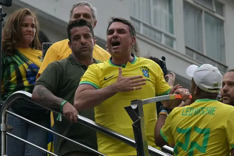 Flávio Bolsonaro veste camisa amarela da seleção brasileira, gesticula com uma mão no peito e aparece cercado por outras pessoas em uma plataforma.