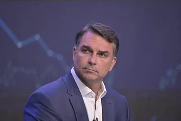 Flávio Bolsonaro, de paletó azul e camisa branca, olha para o lado com expressão séria diante de um painel escuro com linhas azuladas.