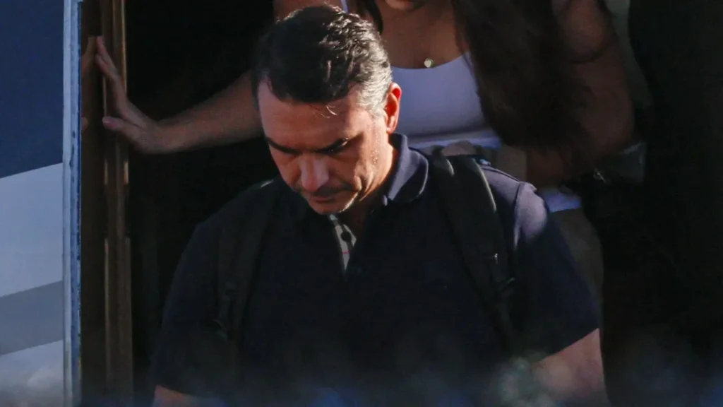 Homem de camisa polo azul-marinho e mochila preta desce pela porta de uma aeronave, olhando para baixo, enquanto outra pessoa aparece ao fundo.