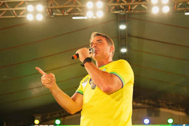 Homem de camiseta amarela com detalhes verdes fala ao microfone e aponta com o dedo indicador sobre um palco iluminado.