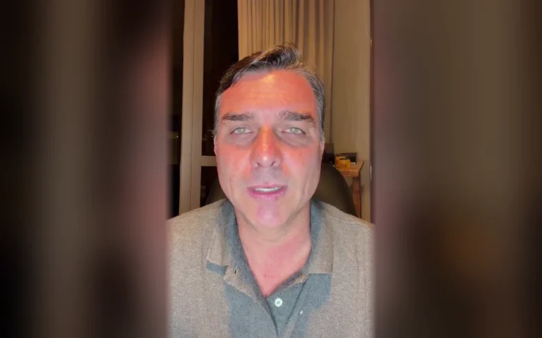 Homem de cabelo grisalho e camisa polo cinza olha para a câmera em ambiente interno, com cortinas ao fundo e laterais desfocadas.