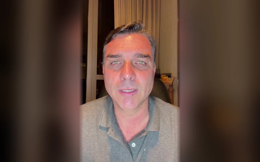 Homem de cabelo grisalho e camisa polo cinza olha para a câmera em ambiente interno, com cortinas ao fundo e laterais desfocadas.