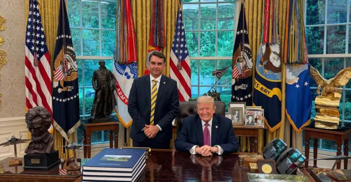 Flávio Bolsonaro em pé ao lado de Donald Trump, sentado à mesa presidencial no Salão Oval da Casa Branca, durante encontro em maio de 2026.