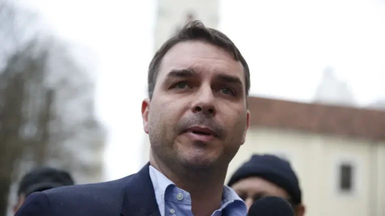 Flávio Bolsonaro, de paletó azul-escuro e camisa azul-clara, fala diante de um microfone, com outras pessoas ao fundo.