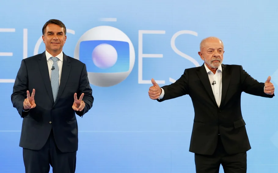 Flávio Bolsonaro faz gestos com as duas mãos enquanto Lula ergue os polegares no palco de debate da Globo.