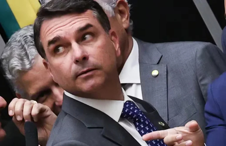 Flávio Bolsonaro, de terno cinza e gravata azul estampada, olha para o lado e gesticula com a mão, com outras pessoas ao fundo.