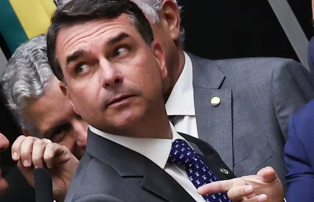 Flávio Bolsonaro, de terno cinza e gravata azul estampada, olha para o lado e gesticula com a mão, com outras pessoas ao fundo.