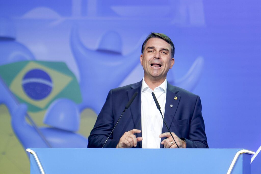 Flávio Bolsonaro usando paletó escuro e camisa branca diante de fundo azul durante convenção do PL.
