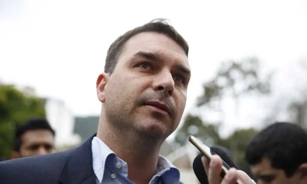 Flávio Bolsonaro, de paletó escuro e camisa azul, fala com jornalistas ao ar livre, diante de um microfone e um celular usado para gravar a entrevista.