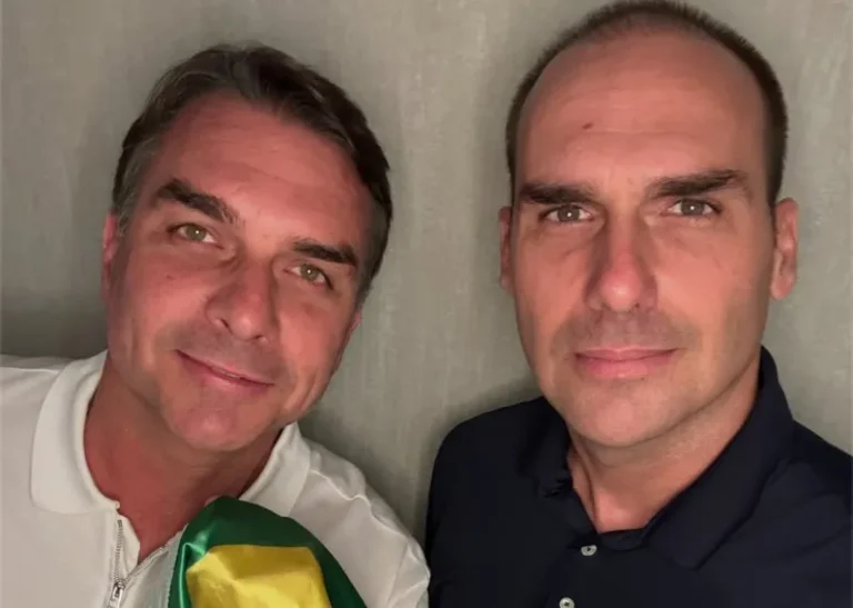 Flávio Bolsonaro e Eduardo Bolsonaro posam juntos nos Estados Unidos, com Flávio segurando uma bandeira do Brasil.