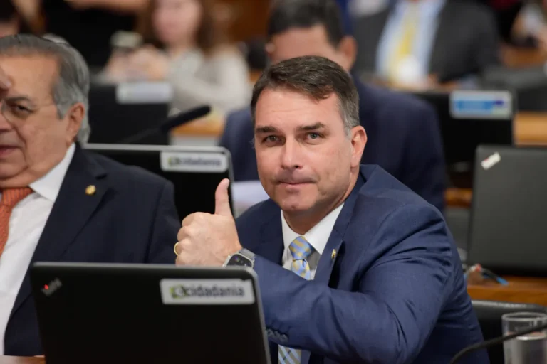 Flávio Bolsonaro, de terno azul e gravata listrada em azul e amarelo, faz sinal de positivo com o polegar enquanto está sentado atrás de um notebook, em ambiente de reunião.