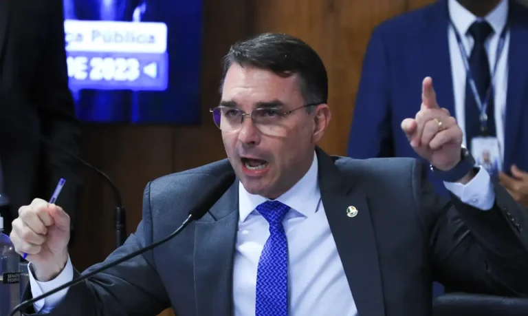Flávio Bolsonaro, de terno escuro e gravata azul, fala ao microfone com o dedo indicador erguido e uma caneta na outra mão.