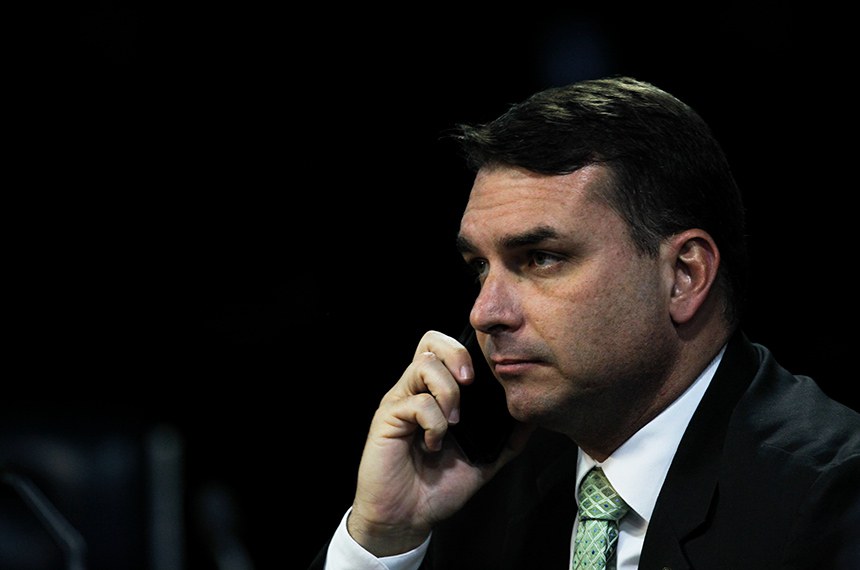 Senador Flávio Bolsonaro falando ao telefone celular, usando terno escuro, camisa branca e gravata verde, com fundo preto.