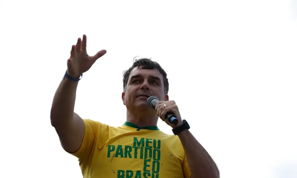 Homem de camiseta amarela com a frase 'Meu partido é o Brasil' segura um microfone e ergue uma das mãos diante de um céu claro.