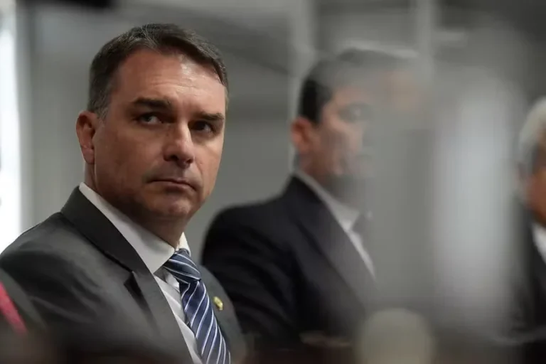 Flávio Bolsonaro veste terno escuro, camisa branca e gravata azul listrada, olhando para o lado, com outras pessoas desfocadas ao fundo.