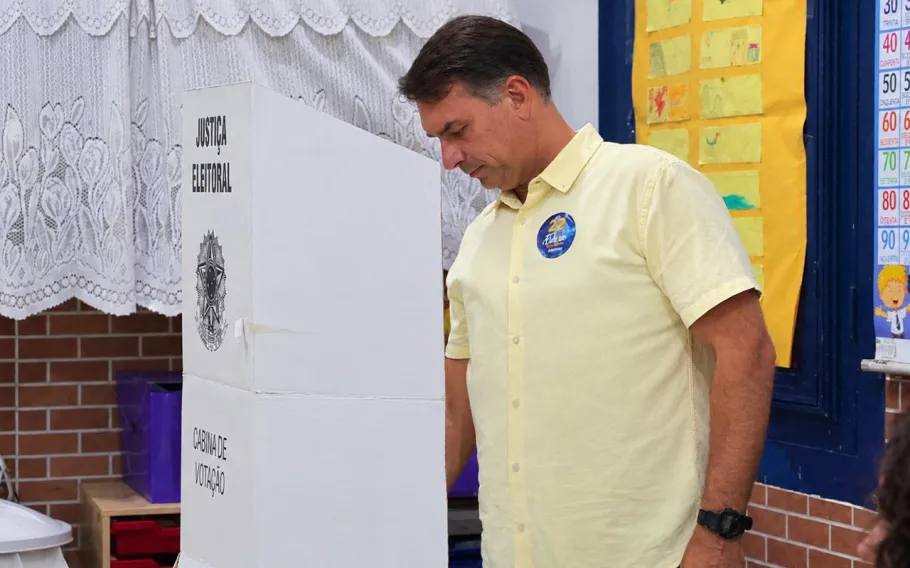 Flávio Bolsonaro, de camisa amarela, diante de uma cabine de votação da Justiça Eleitoral, em sala com cortinas brancas e mural escolar.