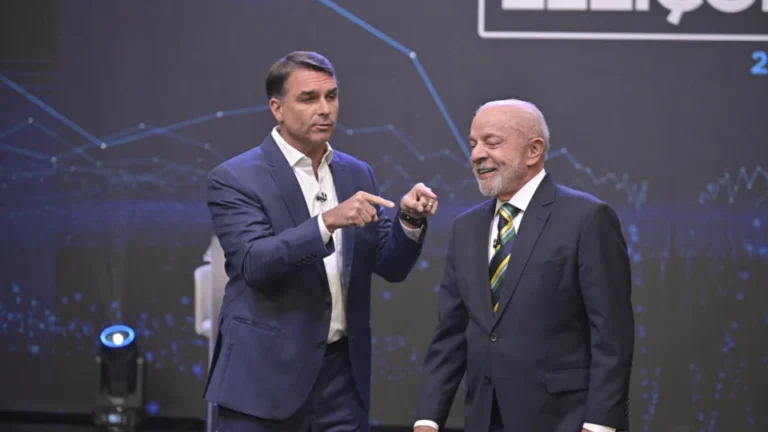 Flávio Bolsonaro, de terno azul, gesticula com as duas mãos enquanto Lula, de terno escuro e gravata verde e amarela, sorri ao seu lado no palco do debate da Band.