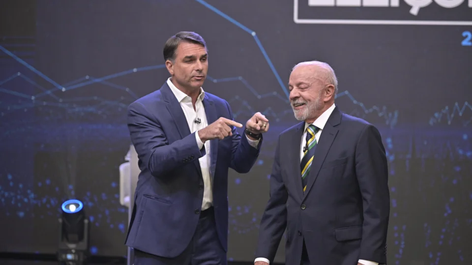Flávio Bolsonaro, de terno azul, gesticula com as duas mãos enquanto Lula, de terno escuro e gravata verde e amarela, sorri ao seu lado no palco do debate da Band.