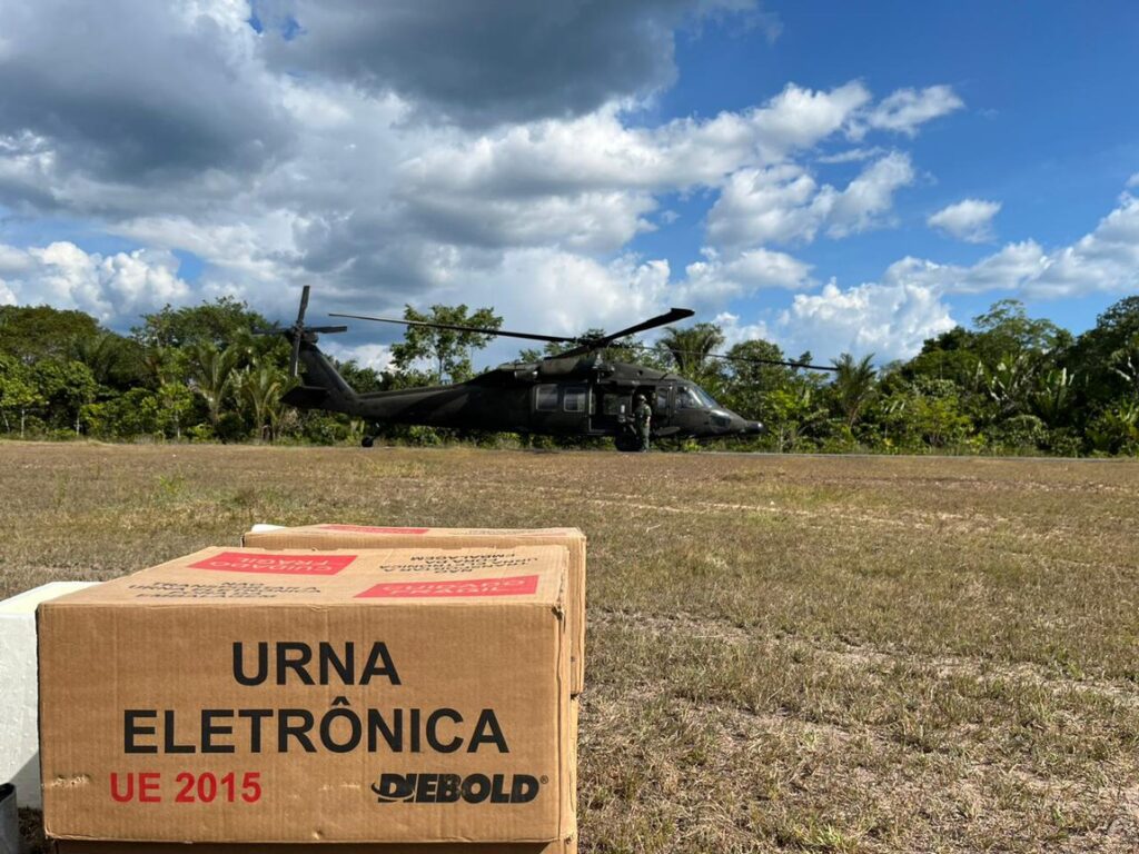 Caixa de urna eletrônica em primeiro plano e helicóptero militar preto pousado em campo aberto, com árvores e céu parcialmente nublado ao fundo.