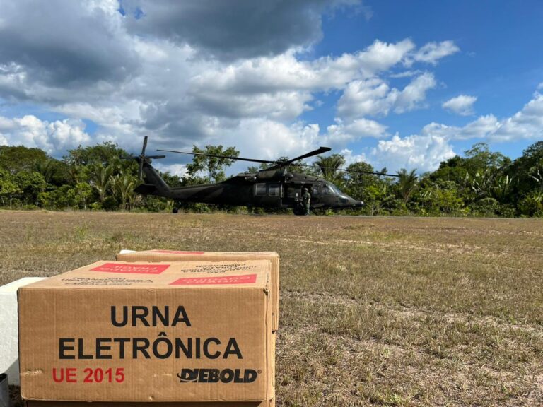 Caixa de urna eletrônica em primeiro plano e helicóptero militar preto pousado em campo aberto, com árvores e céu parcialmente nublado ao fundo.