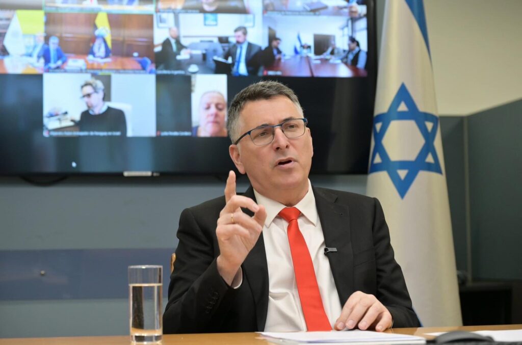 Homem de terno preto e gravata vermelha, com a bandeira de Israel ao fundo, durante videoconferência.
