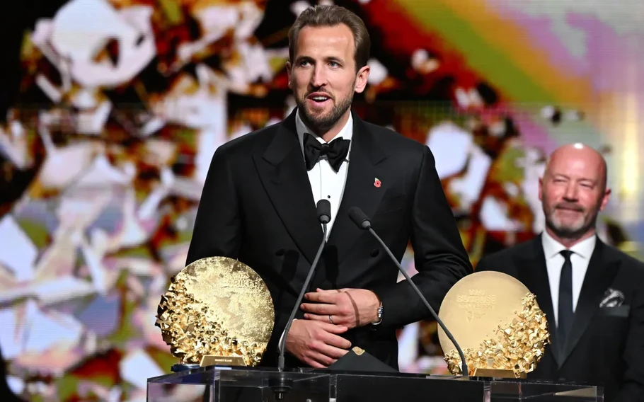 Harry Kane, de smoking preto, fala diante de microfones, com dois troféus dourados à sua frente.