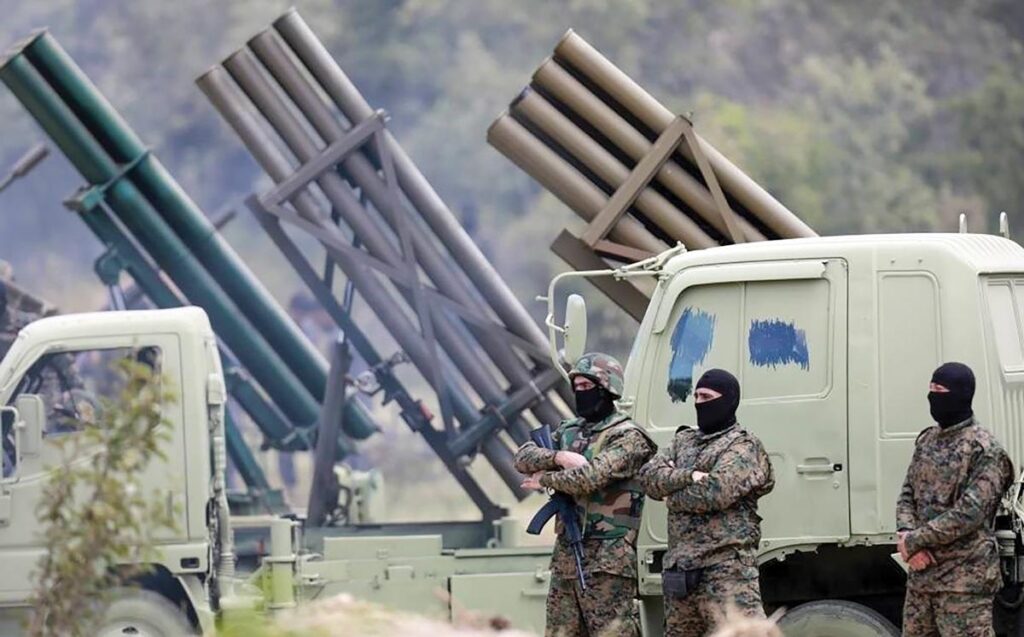 Três integrantes encapuzados do Hezbollah, em uniformes camuflados, diante de caminhões com lançadores de foguetes.