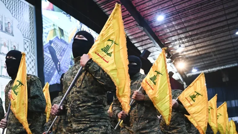 Homens com rostos cobertos e uniformes camuflados seguram bandeiras amarelas do Hezbollah em um evento.