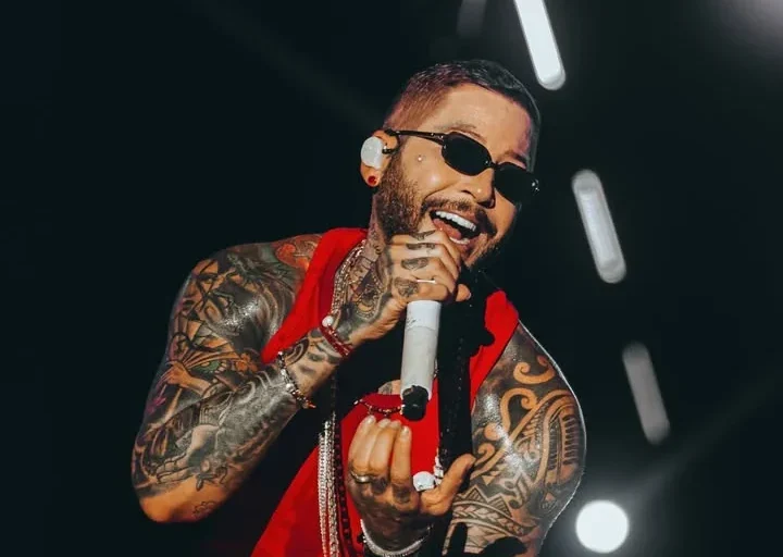Igor Kannário canta ao microfone usando óculos escuros e roupa vermelha, com os braços tatuados e luzes de palco ao fundo.