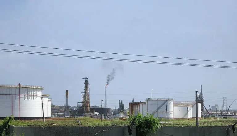 Instalações petrolíferas com grandes tanques brancos de armazenamento, torres industriais e uma chama liberando fumaça.