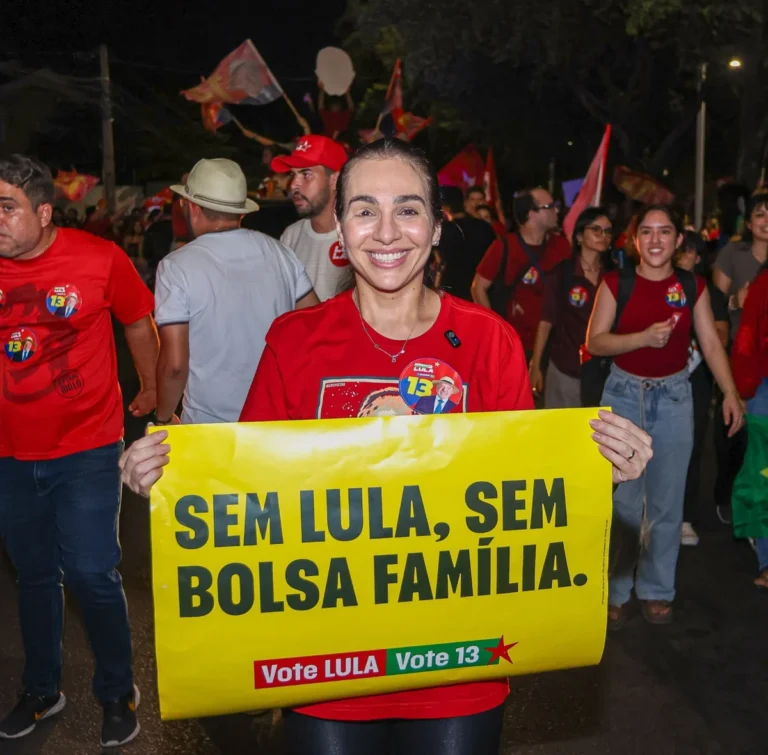 Isabel Fonteles sorri segurando cartaz amarelo com a frase “Sem Lula, sem Bolsa Família” e propaganda eleitoral de Lula, diante de apoiadores vestidos de vermelho.