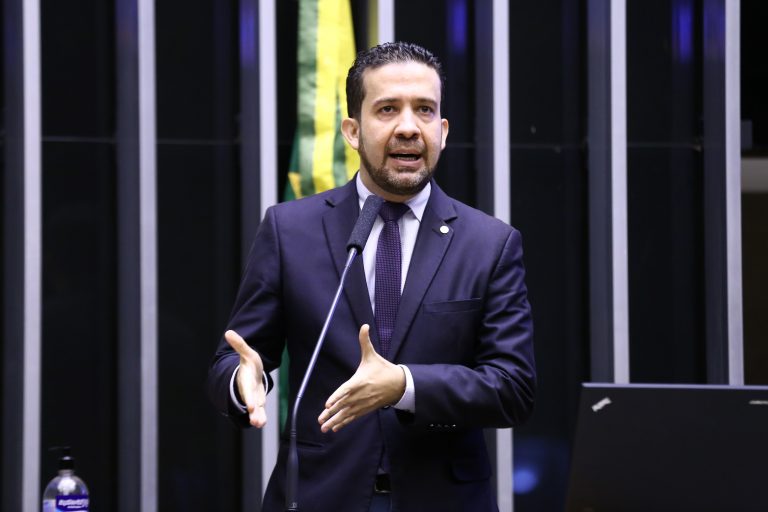 Deputado federal André Janones discursa na tribuna da Câmara dos Deputados, usando terno escuro e gravata, diante de um microfone.