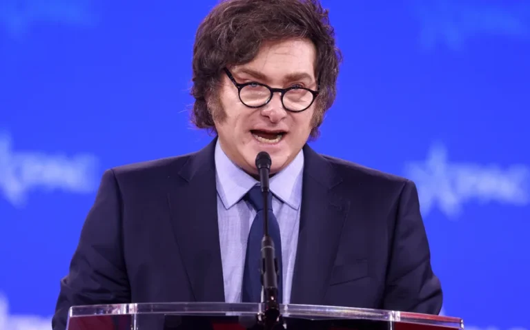 Javier Milei, presidente da Argentina, discursa diante de um microfone, usando terno escuro e óculos, com fundo azul.