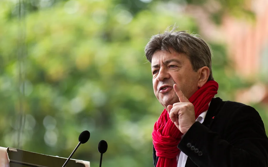 Jean-Luc Mélenchon discursa diante de microfones, usando casaco preto e cachecol vermelho, com o dedo indicador levantado.